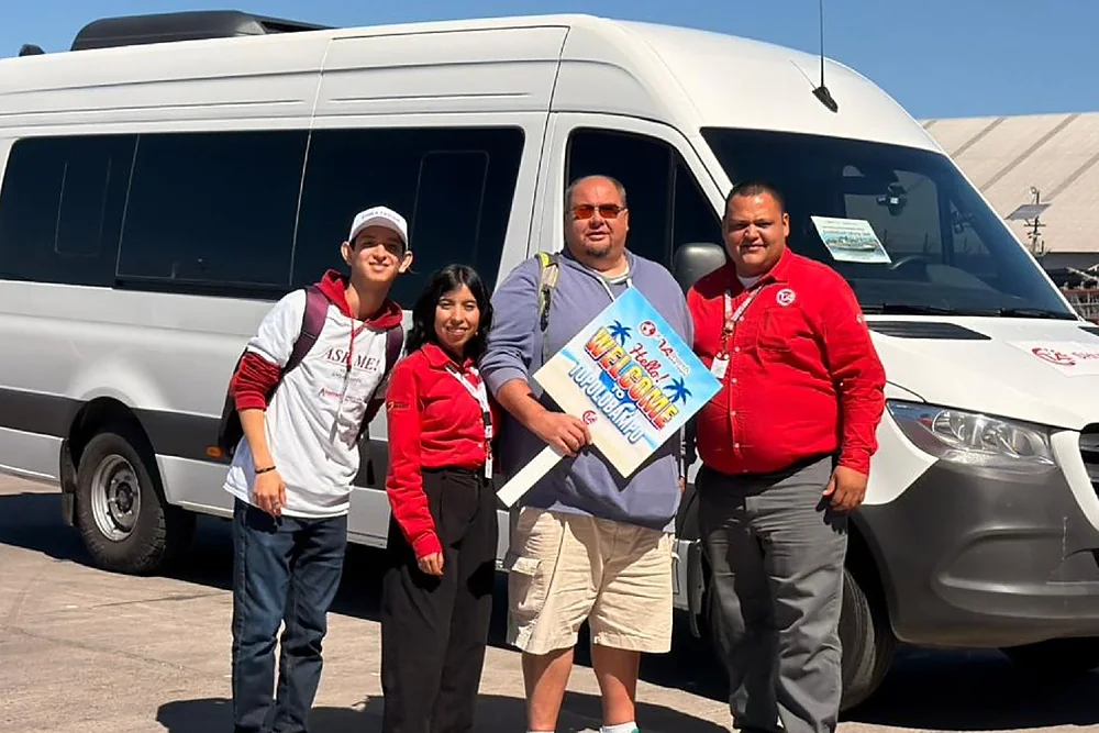 Dos personas del equipo reciben a un pasajero de crucero con un cartel de bienvenida, frente a una van de la agencia